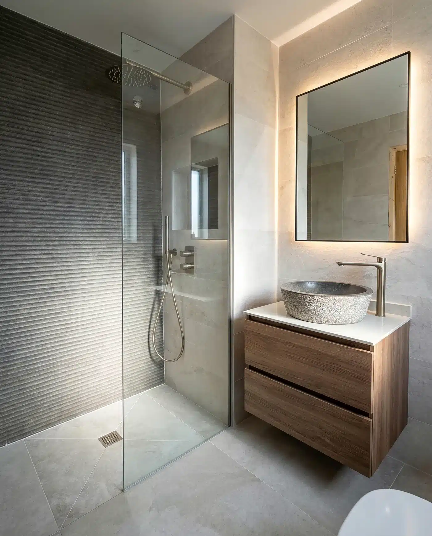 Baño con ducha de mampara fija de cristal, revestimiento gris texturizado y mueble suspendido con lavabo redondo sobre encimera.