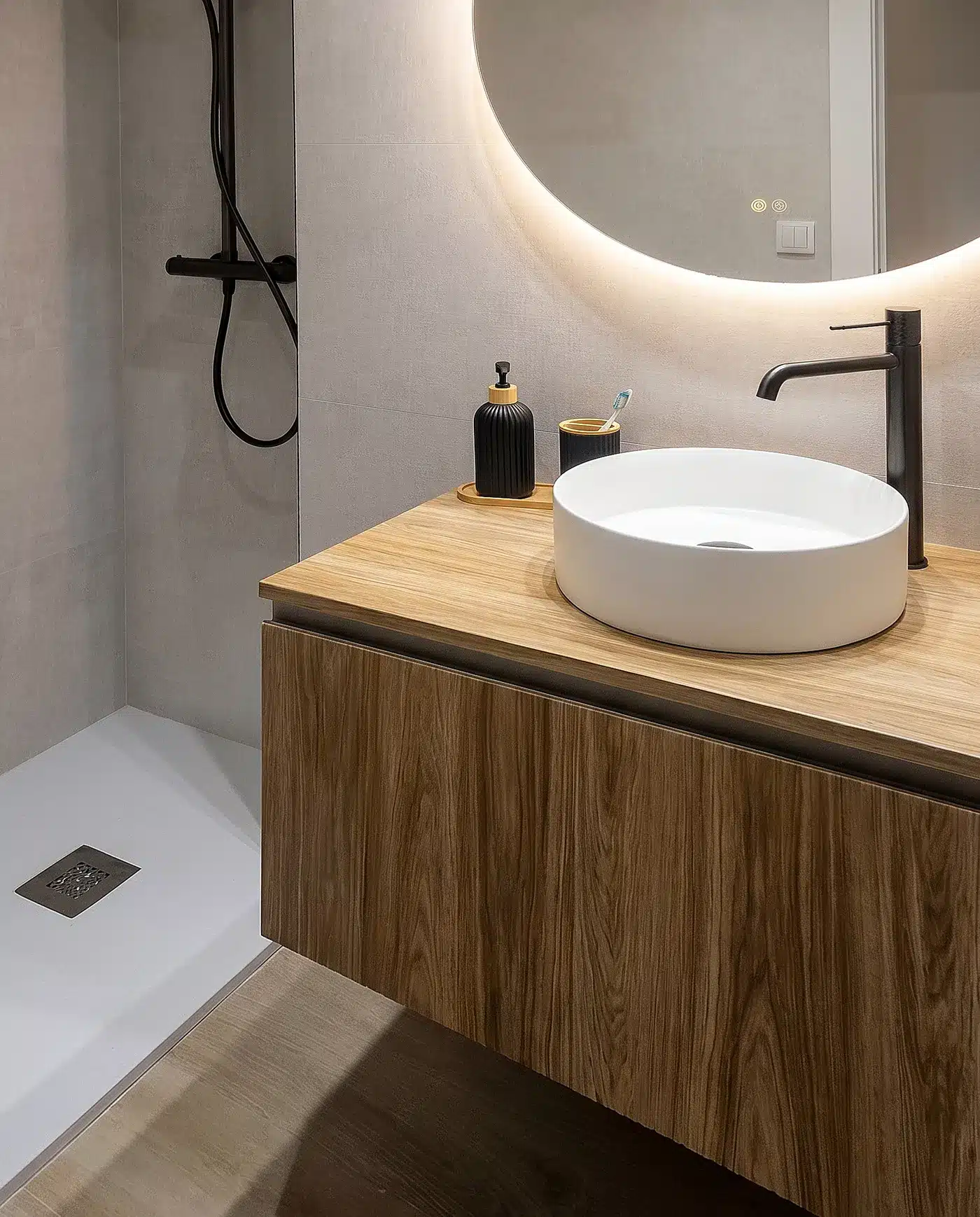 Lavabo blanco sobre mueble suspendido con acabado madera, grifería negra y espejo circular retroiluminado junto a la ducha.