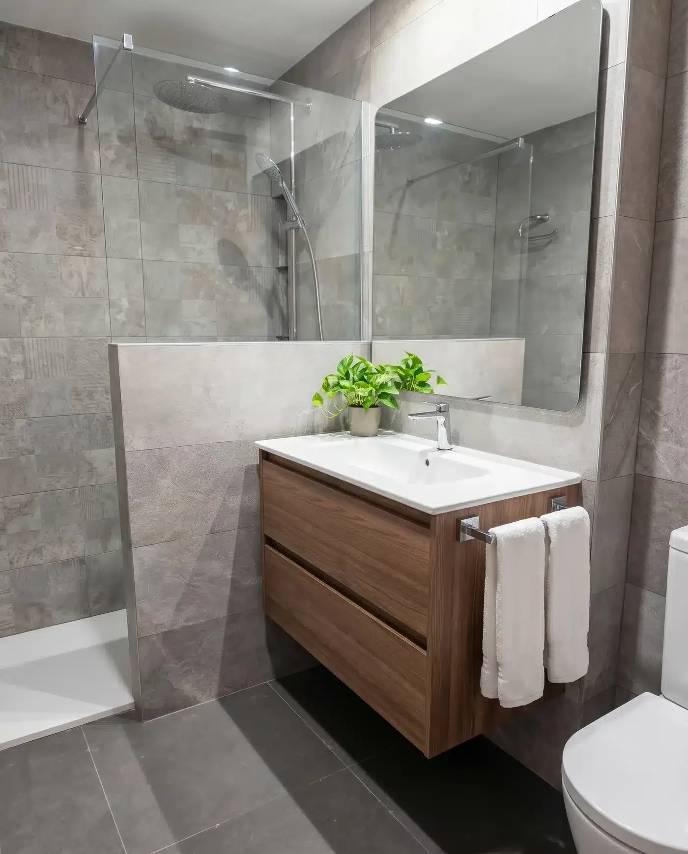 Mueble de baño suspendido con acabado madera, lavabo blanco y toallero lateral junto a una ducha separada por un murete y cristal.
